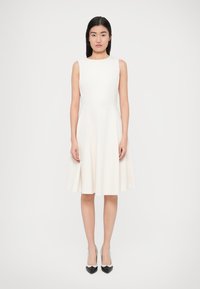 Ένα αμάνικο κρεμ φόρεμα γραμμής Α με εφαρμοστό μπούστο και φουσκωτή φούστα. Φορεμένο με μαύρες και λευκές μυτερές ψηλές γόβες. Λείο υφή.