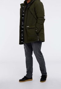 Olive green padded jacket with a zip front and logo detailing, paired with gray jeans and black sneakers featuring a light brown sole.