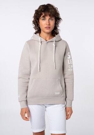 Vrouw met krullend haar die een lichtgrijze hoodie met trekkoorden en witte opgerolde shorts draagt, staand tegen een effen witte achtergrond.