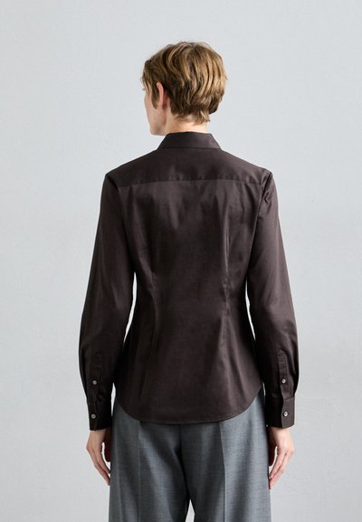Persona in piedi con le spalle alla telecamera, che indossa una camicia a maniche lunghe aderente marrone scuro e pantaloni grigi larghi, di fronte a una parete chiara e uniforme.