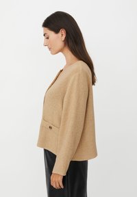 Beige Jacke aus Wollmischung mit weicher Textur, rundem Ausschnitt, Knopfverschluss und einer einzelnen Brusttasche, getragen über einem schwarzen Unterteil.