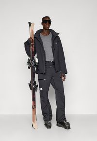 Jachetă neagră de schi cu glugă, cămașă gri, pantaloni negri de schi și bocanci de schi. Ținând schiuri din lemn cu prinderi. Design elegant și detalii funcționale.