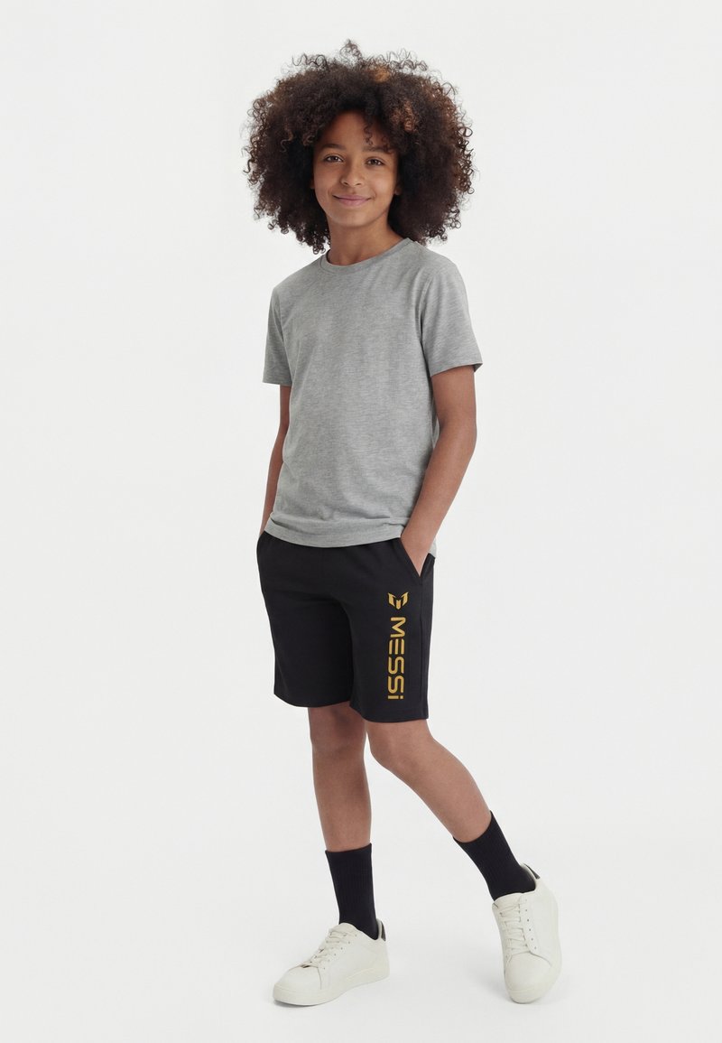 Garçon souriant debout les mains dans les poches portant un T-shirt gris, un short noir Messi, des chaussettes noires et des baskets blanches sur fond blanc.