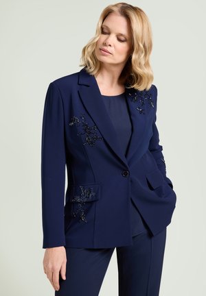 Vrouw in marineblauw pak met kralenbloemborduurwerk op het jasje, staand met één hand in de zak tegen een effen achtergrond.