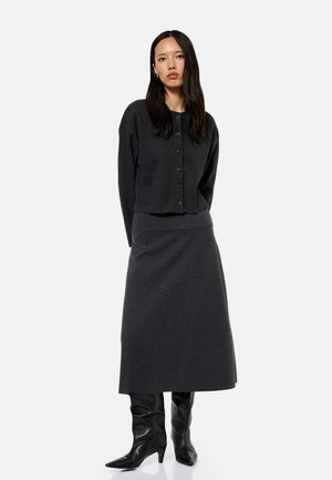 Kobieta ubrana w ciemnoszary sweter zapinany na guziki, dopasowaną midi spódnicę oraz czarne skórzane botki z ostrym noskiem, stojąca na jednolitym tle.