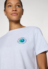 T-shirt en coton blanc avec un graphique circulaire multicolore présentant un motif de vague bleue et du texte. Manches courtes et coupe décontractée.