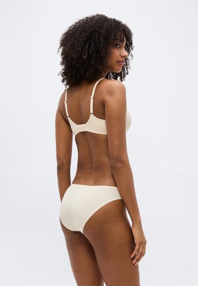 Γυναίκα με σγουρά μαλλιά που φοράει μπεζ σουτιέν και αντίστοιχο εσώρουχο, εμφανιζόμενη από πίσω πάνω σε απλό λευκό φόντο.