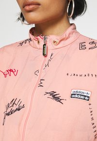 Světle růžová bunda na zip s vyšívaným černým graffiti stylem a nášivkou s logem Adidas. Materiál působí lehce a má texturovaný povrch.