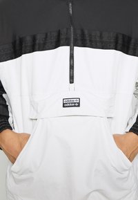 White and black half-zip jacket featuring a black band with embossed Adidas logo, front pocket, and elastic cuffs. Smooth texture, casual design.