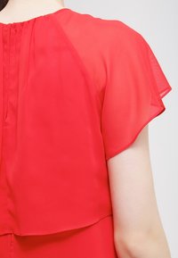 Top en chiffon rouge avec des manches courtes transparentes, design superposé et un détail de plis subtil au niveau de l'encolure. Texture lisse et apparence légère.