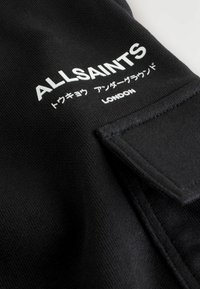 Mustmust kangas, millel on tekstuuriline pind. Valge trükitud tekst "ALLSAINTS" ja "LONDON", millele on lisatud jaapani keelsed tähed. Omab tasku detaili.