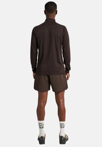 Brun långärmad topp med hög krage, parat med mörka shorts. Modellen bär vita strumpor med svart text och gråskor.