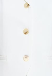 Tissu blanc avec trois boutons beige, montrant une boutonnière et une poche latérale partielle sur le devant d'un vêtement.