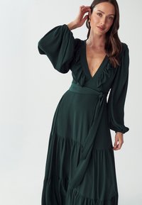 Long green dress made of textured fabric, featuring a deep V-neck, ruffled accents, and billowy sleeves with a waist tie and tiered skirt.