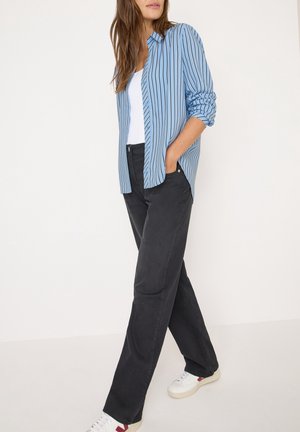 Vrouw draagt een blauw gestreept overhemd met knopen over een wit topje, zwarte rechte spijkerbroek en witte sneakers, staand met één hand in de zak.