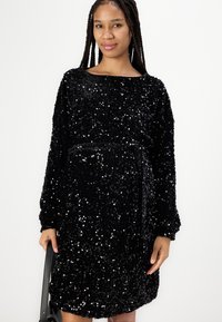 Vestido preto de lantejoulas com mangas longas, decote redondo e cintura cingida. Apresenta uma textura suave e um ajuste descontraído.