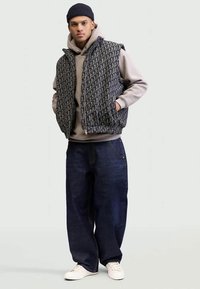 Jovem usando um gorro preto, colete com padrão preto e cinza com fecho zip, moletom bege, calças largas azul-escuro e ténis brancos, de pé com as mãos nos bolsos.