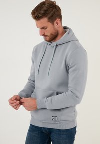 Sudadera con capucha gris, con un bolsillo frontal, cordón y puños acanalados. La etiqueta en la parte inferior izquierda presenta un logo. Tela suave y texturizada.