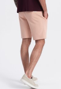 Shorts rose clair avec des poches latérales et une texture douce, associés à des baskets beiges. Le modèle porte une chemise bordeaux foncé.