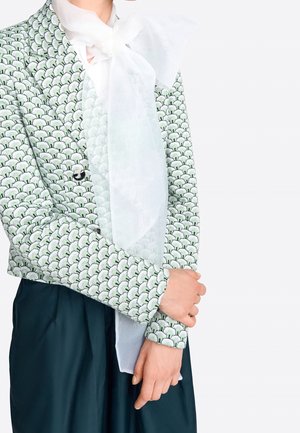 Personne portant une veste verte et blanche à motif avec un grand nœud blanc transparent noué au cou, et un pantalon ample de couleur foncée.