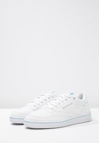 Sneakers in tela bianca con design basso, dettagli blu lungo le suole, superficie testurizzata e chiusura semplice con lacci.