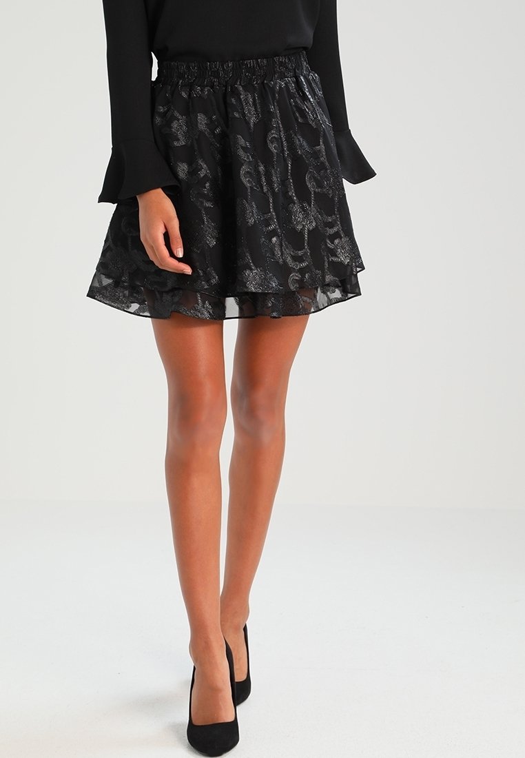 Femme portant un haut à manches longues noir, une jupe courte noire à motifs et des chaussures à talons noirs, debout avec une main près de la jupe.