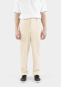 Beige veckade byxor med en avslappnad passform, tillverkade av lättviktsmaterial. Har en midjevecka med en knapp och sidofickor, kombinerat med svarta sneakers.