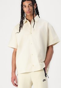 Μισοαντίθετος, oversized πουκάμισο με κουμπιά και κοντό μανίκι σε ανοιχτό κίτρινο βαμβάκι. Έχει ένα μικρό λευκό λογότυπο Nike στο στήθος και χαλαρή εφαρμογή.