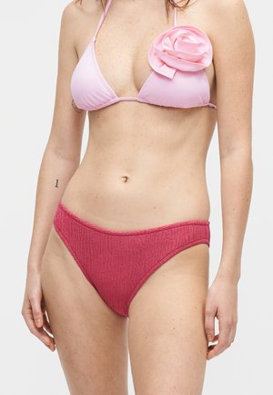 Γυναίκα που φοράει τοπ ροζ μπικίνι με λεπτομέρεια ένα τριαντάφυλλο και υφασμάτινο σκούρο ροζ κάτω μέρος μπικίνι, στέκεται μπροστά σε ένα μονόχρωμο ανοιχτό φόντο.