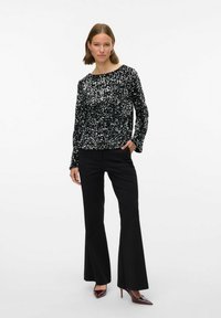 Črni bleščeči top z dolgimi rokavi, okroglega izreza in ohlapnim krojem, v kombinaciji s črnimi širokimi hlačami in čevlji z ostro konico.