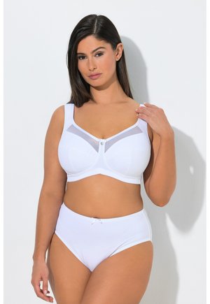 Mujer con un sujetador blanco de cobertura completa con detalle de malla y bragas de talle alto a juego, de pie contra un fondo claro y liso.