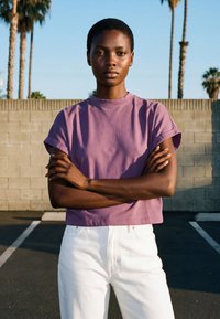 Persona joven de pie al aire libre con los brazos cruzados, vistiendo una camiseta morada de manga corta y pantalones blancos, frente a una pared y con palmeras de fondo.