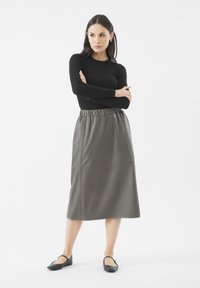Langer, olivgrüner Lederrock mit einem elastischen Bund, kombiniert mit einem schwarzen Langarmtop und flachen schwarzen Schuhen, vor einem weißen Hintergrund.