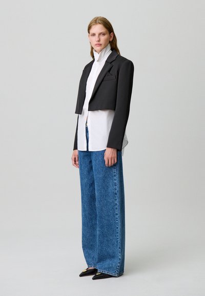 Blazer negro corto sobre una camisa blanca con cuello, combinado con jeans azules holgados y zapatos de punta negra. Diseño simple y ajuste a medida.
