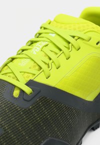 Φωτεινό κίτρινο αθλητικό παπούτσι με επάνω μέρος από δίχτυ, ενισχυμένη μύτη και υφή σε γκρι λεπτομέρειες. Διαθέτει κορδόνια και σόλα με σχέδιο για καλύτερη πρόσφυση.