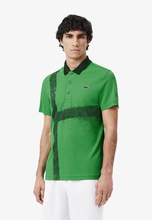 Camisa polo verde con cuello negro, que presenta un patrón de salpicaduras negras diagonales en la parte delantera y un pequeño logo en el pecho.