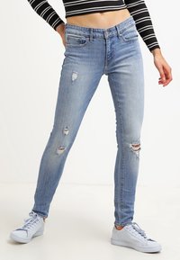 Light blue denim skinny jeans with distressed detailing, featuring several rips and a mid-rise waist. Paired with white sneakers.