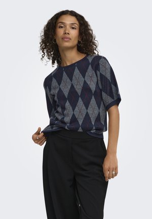 Femme aux cheveux bouclés portant un pull à manches courtes bleu marine et gris à motif losanges et un pantalon noir, debout devant un fond uni.
