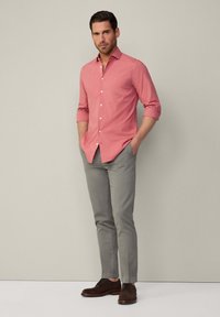 Camisa de botón color coral claro con mangas remangadas, combinada con pantalones ceñidos gris claro y zapatos marrones oscuros. Fondo neutro.