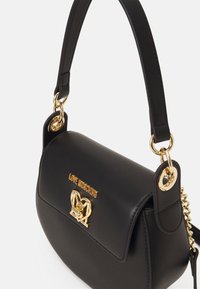 Sac à main en simili cuir noir avec une forme structurée, quincaillerie dorée et un logo en forme de cœur à l'avant. Comprend une bandoulière en chaîne amovible.