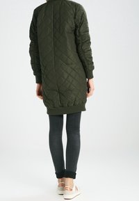 Long quilted olive green coat with a round neckline, ribbed cuffs, and hem; paired with dark jeans and white sneakers.