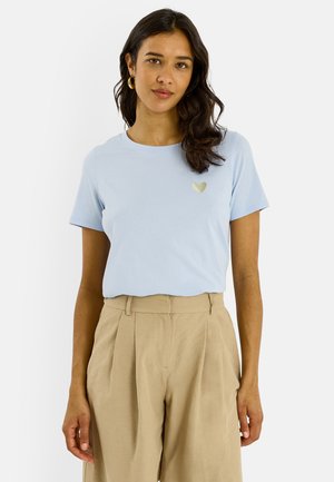 Femme aux cheveux ondulés foncés porte un T-shirt bleu clair avec un petit cœur doré sur la poitrine gauche et un pantalon plissé beige taille haute.