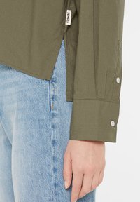 Olive green long-sleeve shirt with a rounded hem, buttoned cuffs, and side slits, paired with light blue denim jeans.