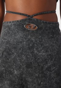 Schwarzes Stretch-Top aus Stoff mit texturierten Details, gekreuzten Trägern und einem auffälligen silbernen Logo-Highlight.