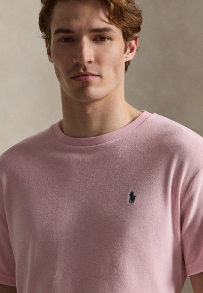 Νεαρός άνδρας με ανοιχτό καστανό μαλλί που φοράει ένα ανοιχτό ροζ μπλουζάκι με ένα μικρό κεντημένο σκοτεινό λογότυπο στο στήθος, μπροστά σε ουδέτερο φόντο.