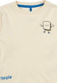 Krēmkrāsas T-kreklis ar īsām piedurknēm, uz krūšu zonas ir izšūta kartonu maizes šķēle un apkakles malā ir zila šuvuma detaļa.