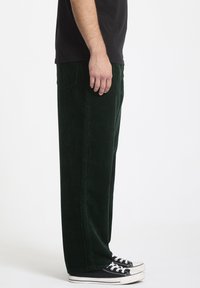 Groene corduroy broek met brede pijpen, voorzien van getextureerde verticale ribbels. Gepaard met zwarte sneakers, staande tegen een effen achtergrond.