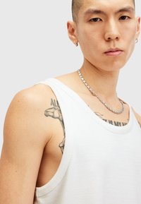 Witte katoenen tanktop met brede armsgaten. Kenmerkt zich door een gestructureerde stof, samengevoegd met een zilveren kettinghalsband en zichtbare tatoeages op de arm.