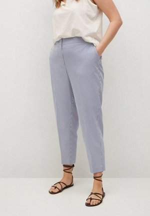 Vrouw draagt lichtblauwe en witte fijn gestreepte broek met zakken, een los wit mouwloos topje en zwarte platte sandalen met bandjes.