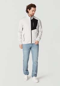 Vit zip-up jacka med svart accent på bröstet, två sidofickor, matchad med ljusblå jeans och vita sneakers. Slätt material.